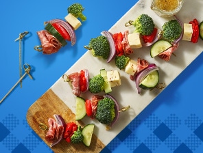 Salami, cubed cheese, broccoli, red pepper and other vegetables on skewers, with dressing on the side. Served alongside a bowl of potato chips.