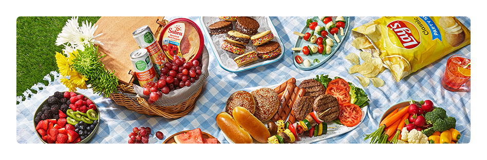 Bowl of sliced fruit, platter of grilled hotdogs and hamburgers, paper-wrapped sandwiches, and more fresh summer foods assorted on a picnic blanket.