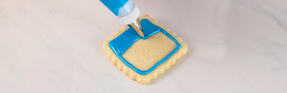 A square sugar cookie on a white mar-ble surface. Piping tip is applying a base layer of blue royal icing.