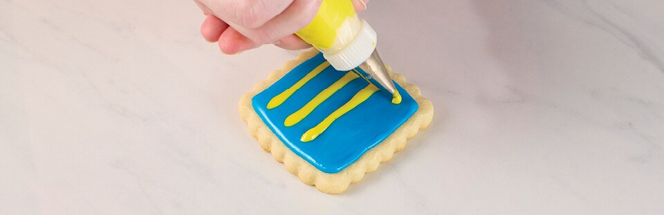 A square sugar cookie on a white marble surface with blue base icing. Piping tip is applying a series of yellow icing lines overtop.