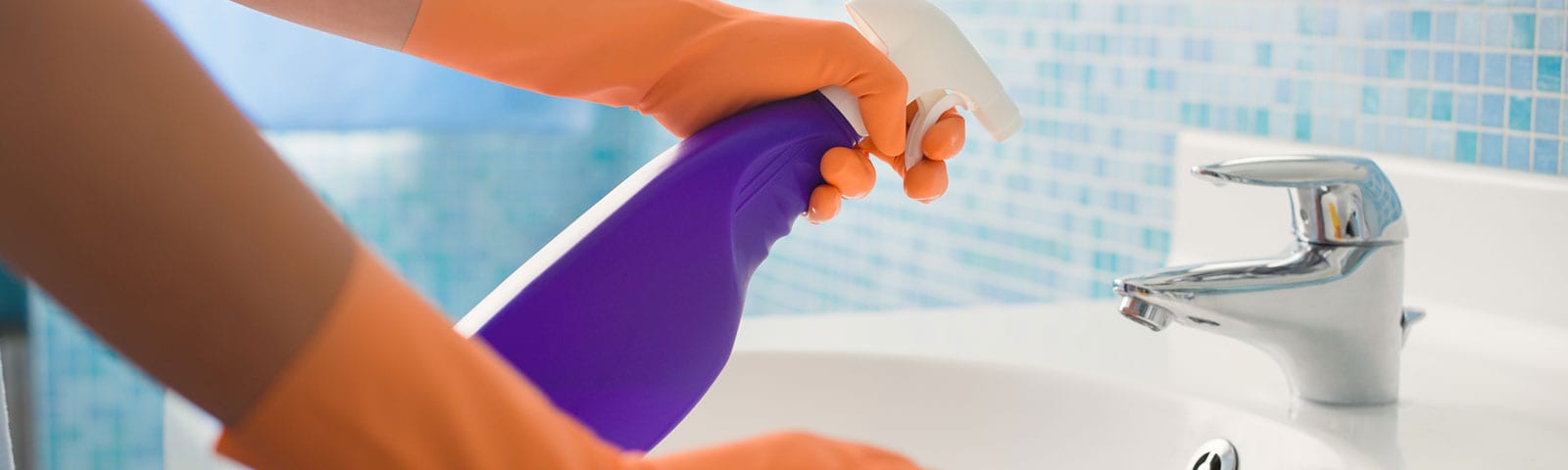 Two arms wearing rubber gloves cleaning a bathroom sink, spray bottle in the left hand with a cloth to wipe in the right