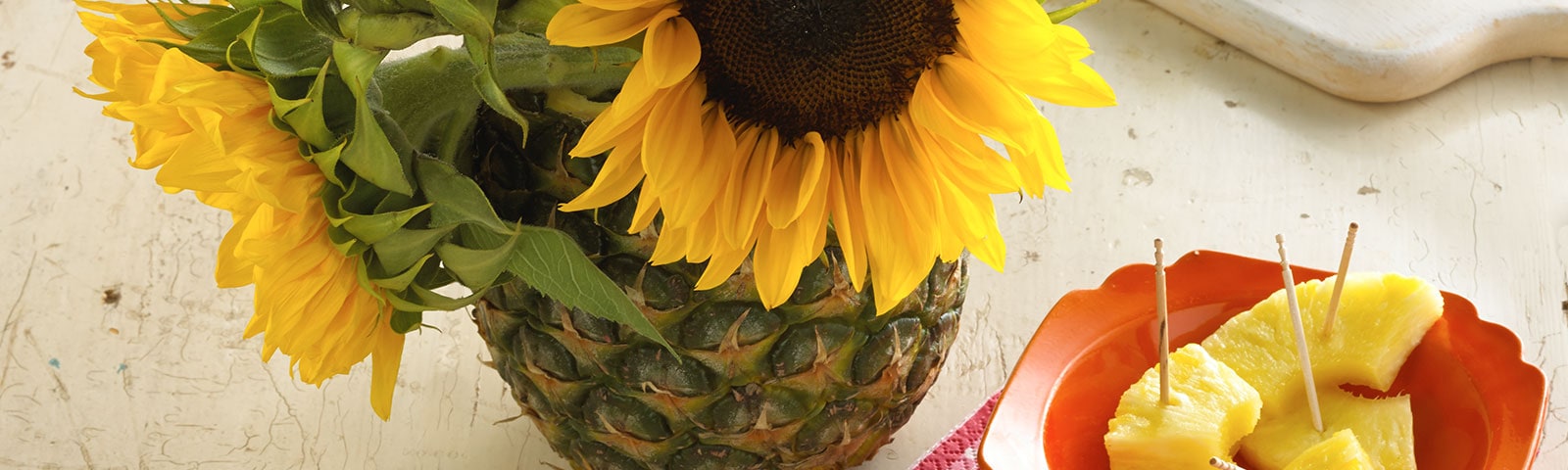 Close crop from above of a pineapple turned into a vase with large sunflowers sitting inside. To the right a plate with pineapple chunks. Close crop from above of a pineapple turned into a vase with large sunflowers sitting inside. To the right a plate with pineapple chunks.