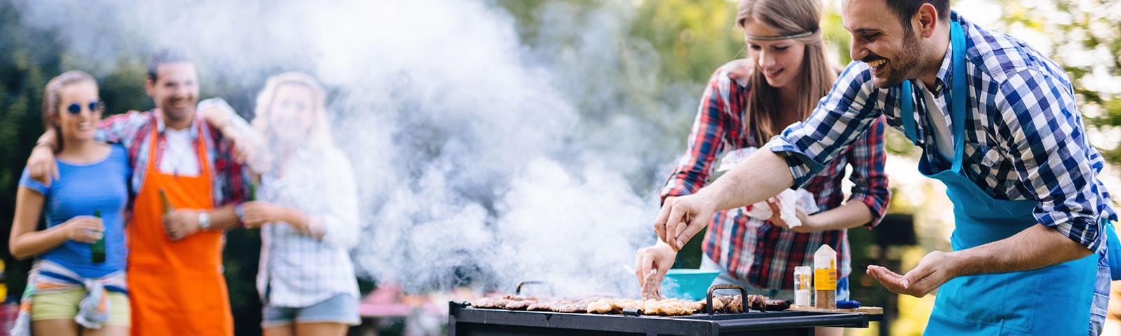 Outdoor grill with two people standing over it turning the hot dogs. In the background a group of friends watches on. Outdoor grill with two people standing over it turning the hot dogs. In the background a group of friends watches on.