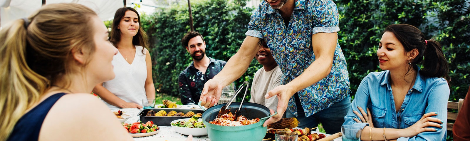 Party guests seated around a table smiling while a man lowers a bowl of food onto the center of the table. Party guests seated around a table smiling while a man lowers a bowl of food onto the center of the table.